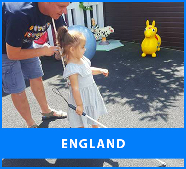 England. Image: Lead Visioneer Daniel Kish bends over as he instructs a young blind girl on using her long Perception Navigation Cane on the patio in her back yard on a sunny day.