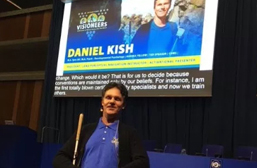 Daniel Kish presents a Keynote Address at the ZeroCon 2018 event at the United Nations Headquarters in Vienna, Austria. Image: Daniel stands in the foreground while a raised screen shows a live video screen of his address as well as a large photo slide and real-time captioning.