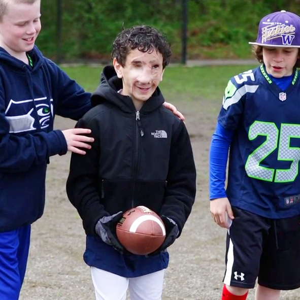 Image: Video still frame from 2014 shows an 11 year-old Humoody Smith holding a football in between two of his teammates as one of them guides him by the shoulders into the direction the ball needs to go.